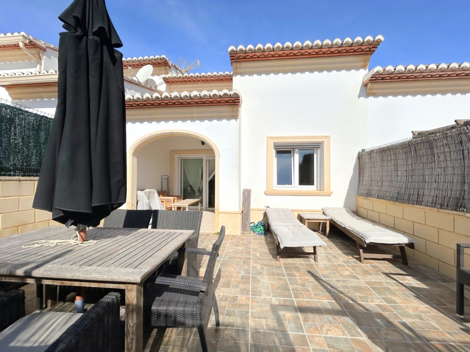 Terrasse privée avec sol en grès, espace repas extérieur et chaises longues. La façade méditerranéenne dispose d'un porche voûté et d'une toiture en tuiles classiques.