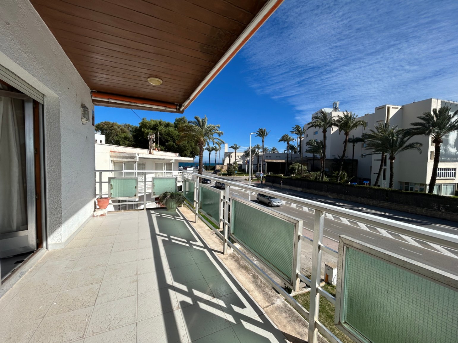 Terrasse extérieure avec sol en céramique, plafond lambrissé en bois et garde-corps métallique. Elle offre une vue dégagée sur l'avenue bordée de palmiers.