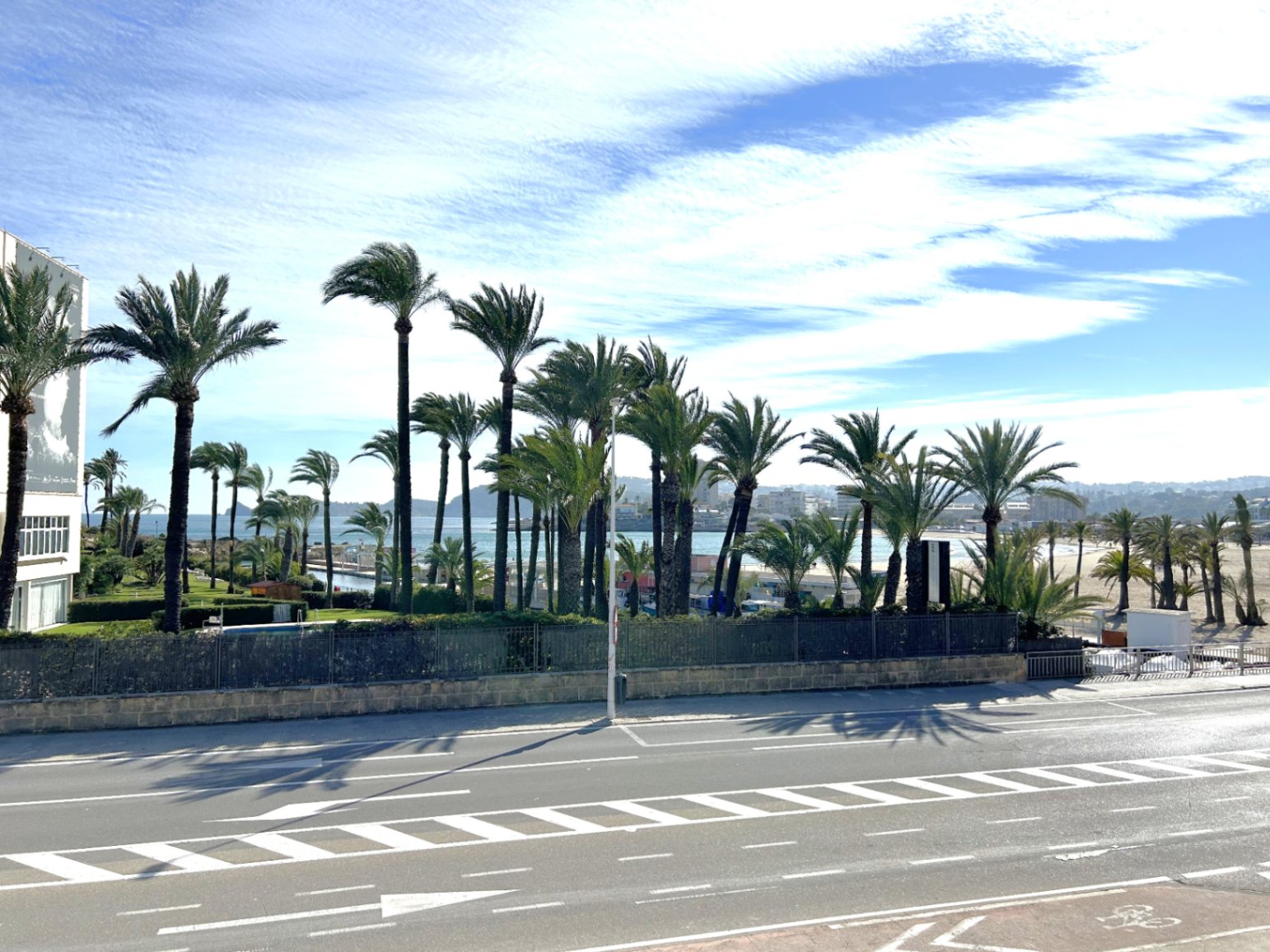 Vue surélevée depuis la terrasse donnant sur une route côtière, une rangée de palmiers et la mer Méditerranée en arrière-plan.