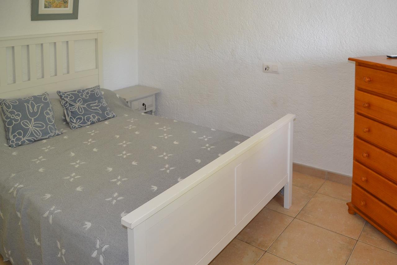 Chambre à coucher avec cadre de lit en bois blanc, sol en grès cérame et commode en bois naturel. Les murs blancs texturés maximisent la clarté de la pièce.