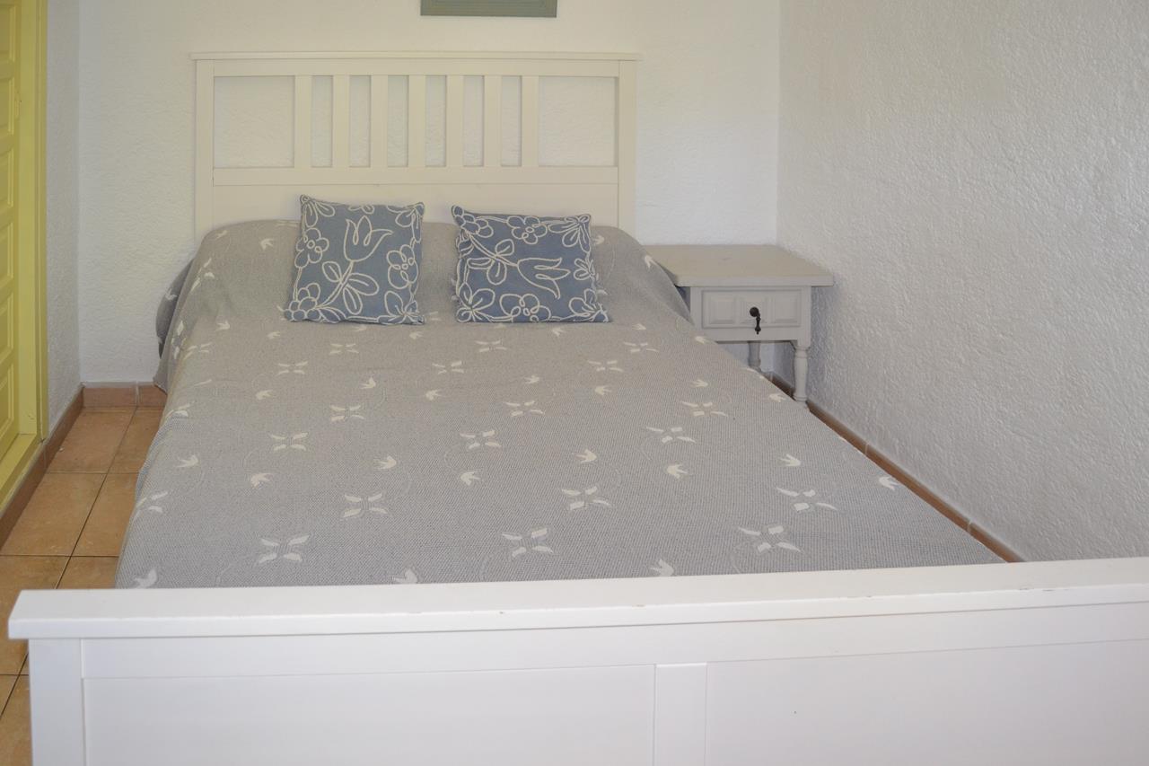 Chambre à coucher avec lit double en bois blanc, table de chevet assortie et sol en grès. Murs blancs texturés et linge de lit aux motifs gris et bleus.