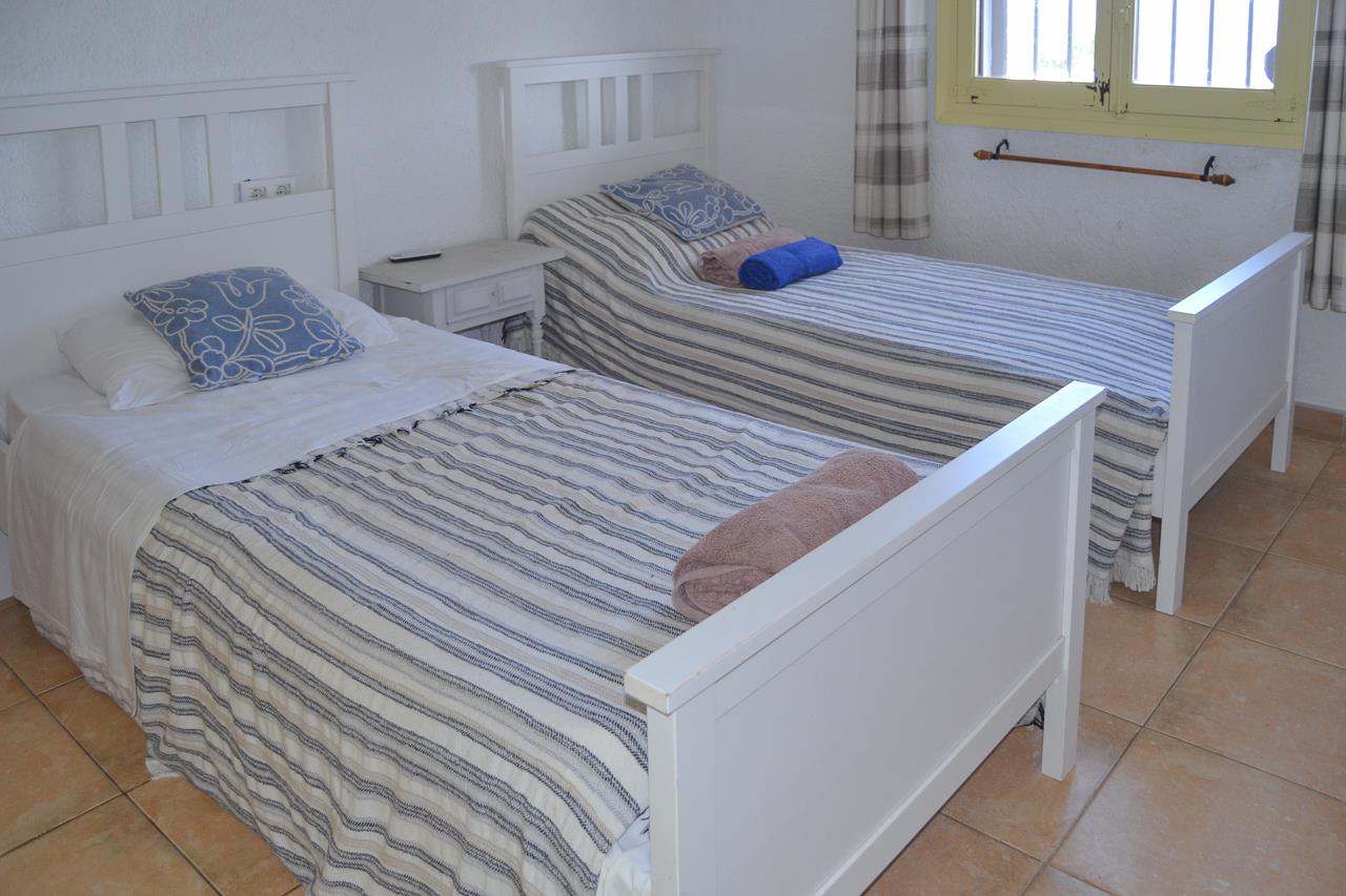 Chambre avec deux lits simples en bois blanc, sol en carrelage et fenêtre équipée de grilles de sécurité et de rideaux à carreaux.