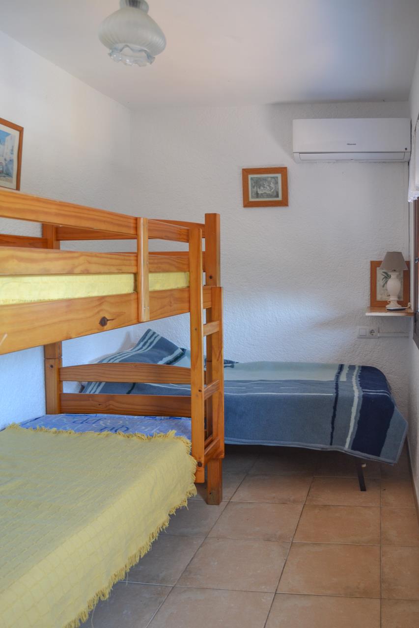 Chambre équipée de lits superposés en pin et d'un lit simple, avec climatisation murale, sol en grès cérame et murs à crépi blanc.