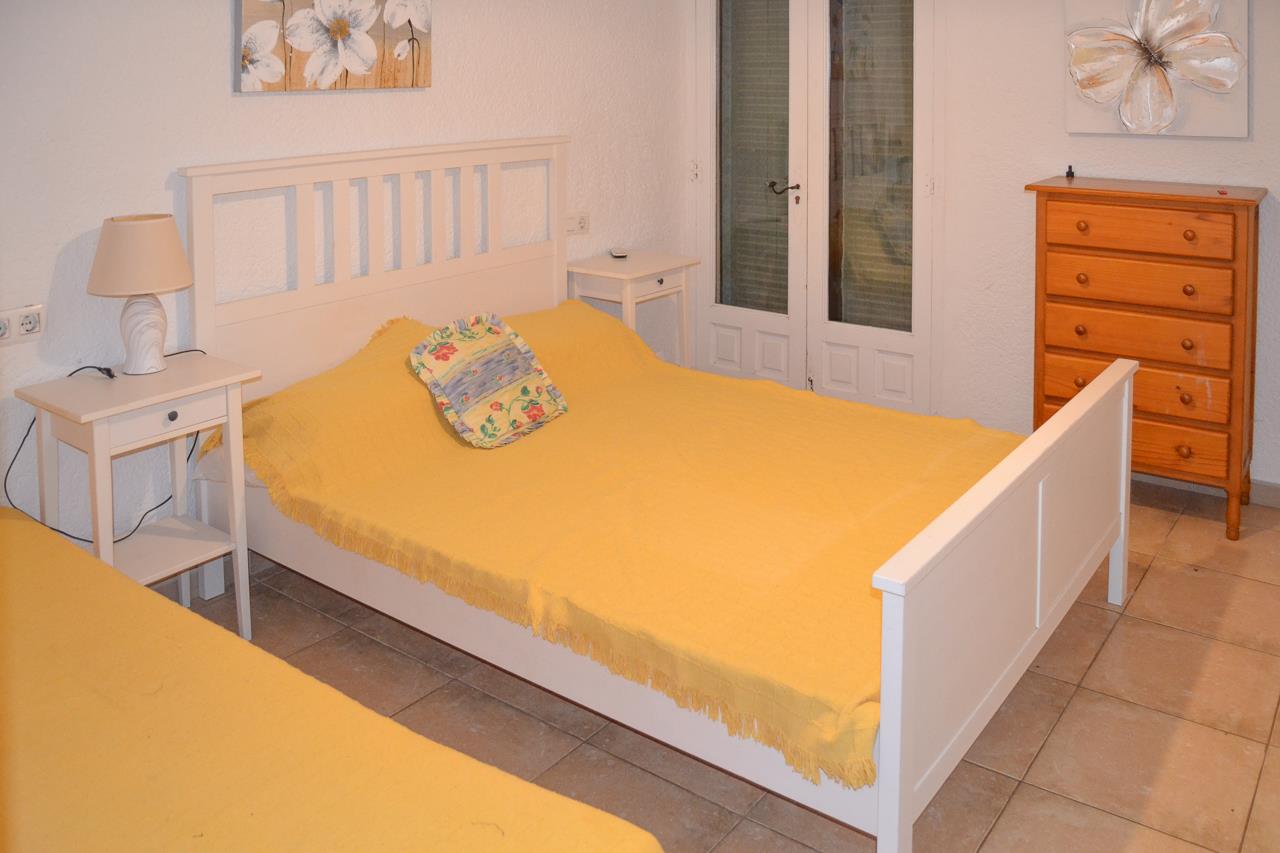 Chambre lumineuse avec cadre de lit en bois blanc, tables de chevet assorties et commode en bois naturel. Sols en carrelage et accès direct vers l'extérieur.