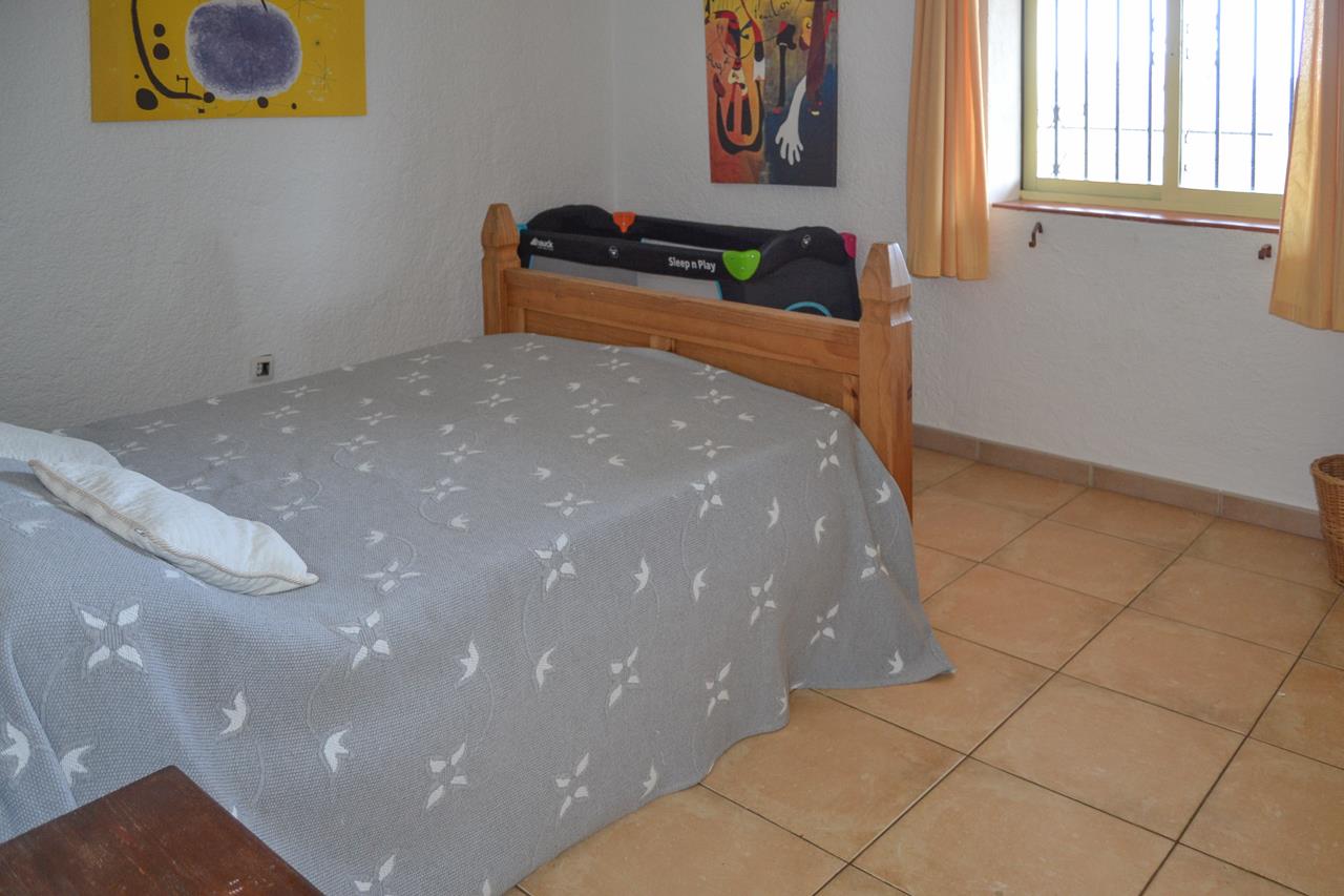 Chambre lumineuse avec lit double en bois, lit parapluie et sol en grès cérame. Elle dispose d'une fenêtre avec barreaux, rideaux jaunes et tableaux décoratifs.