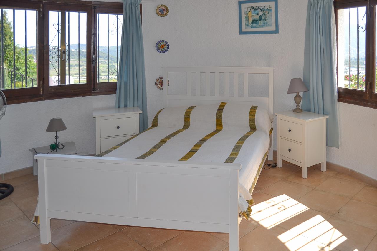 Chambre lumineuse avec lit en bois blanc, sol en terre cuite et fenêtres à cadres en bois sombre offrant une vue dégagée sur la vallée environnante.