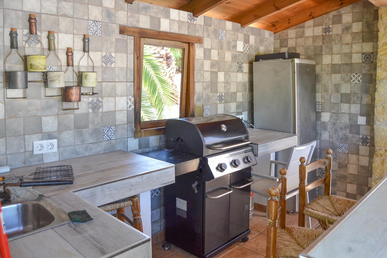 Cuisine d'été couverte avec barbecue à gaz, plans de travail carrelés et murs en mosaïque rustique. Équipée de poutres en bois et d'un évier, avec vue sur le jardin.