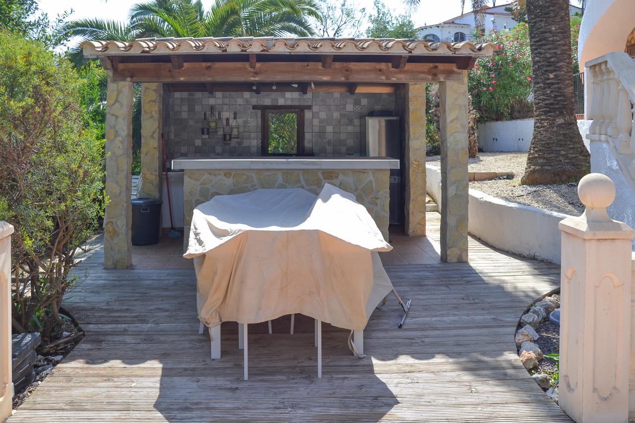 Cuisine d'été extérieure avec bar en pierre, piliers en maçonnerie et toiture en bois sur terrasse en bois, entourée d'un jardin méditerranéen.