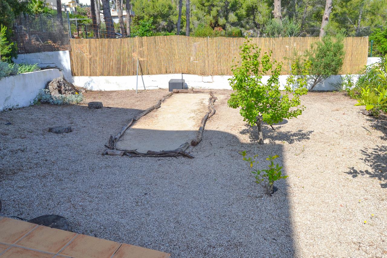Espace jardin en gravier comprenant un terrain de pétanque délimité par des rondins de bois, entouré de végétation méditerranéenne et d'une clôture en canisse.