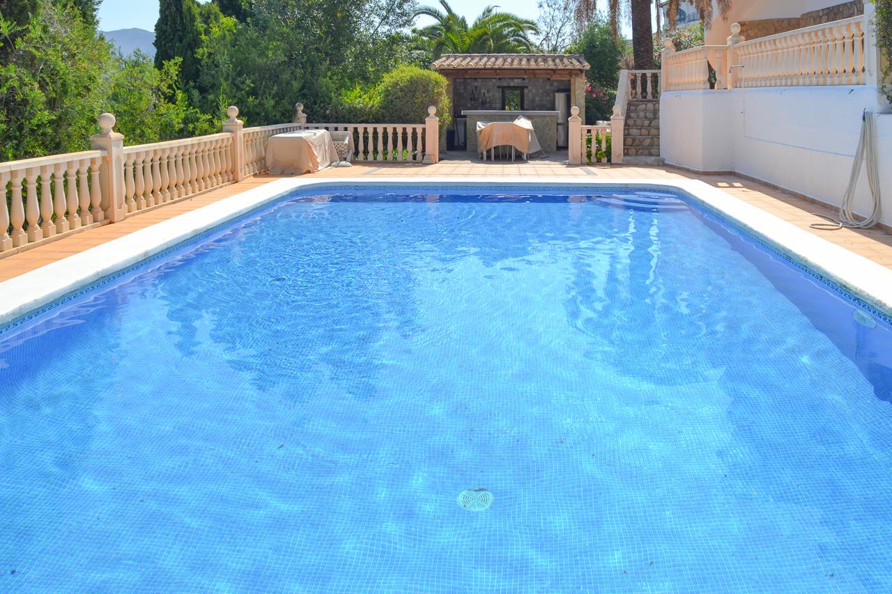 Grande piscine de 12x6 mètres avec mosaïque bleue, entourée d'une terrasse en terre cuite avec balustrades et d'une cuisine d'été couverte.