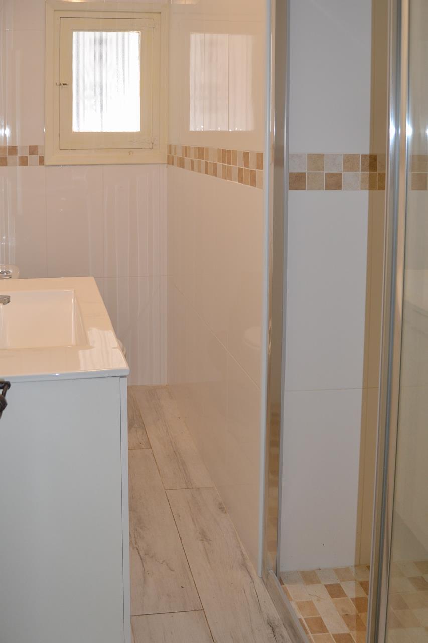Salle d'eau fonctionnelle avec meuble vasque blanc, sol en grès cérame aspect bois et douche vitrée ornée d'une frise en mosaïque.
