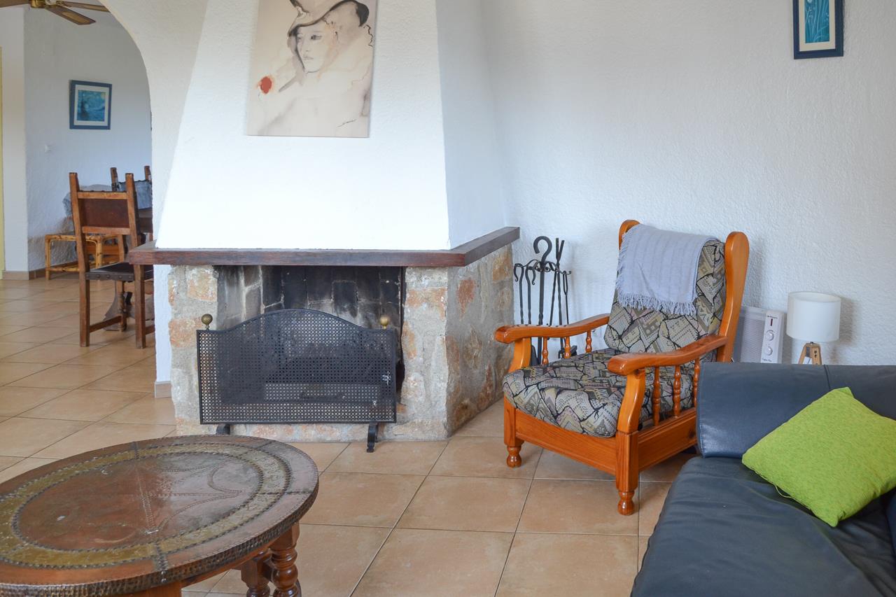 Salon avec cheminée rustique en pierre et bois, carrelage au sol et fauteuil confortable. Une arche s'ouvre sur la salle à manger attenante.