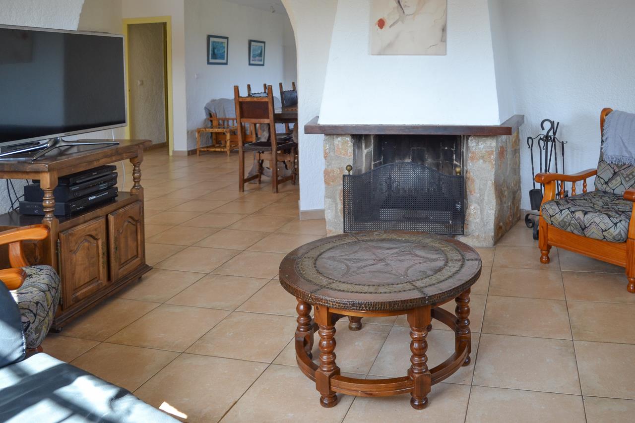 Salon rustique avec cheminée en pierre et hotte blanche, sol en grès cérame et mobilier en bois traditionnel s'ouvrant sur la salle à manger.