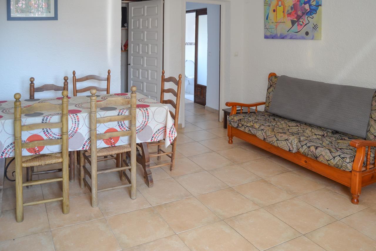 Salon-salle à manger avec sol en grès cérame, table en bois et chaises en paille, ainsi qu'un canapé rustique. La pièce s'ouvre sur un couloir menant à la salle de bain.