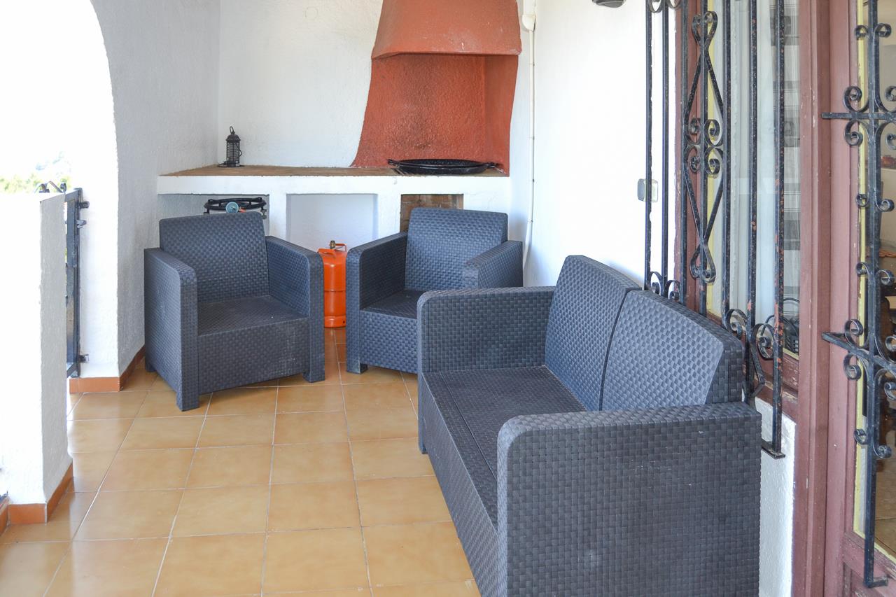 Terrasse couverte avec salon de jardin en résine tressée foncée, sol en terre cuite et barbecue maçonné avec hotte de couleur ocre.