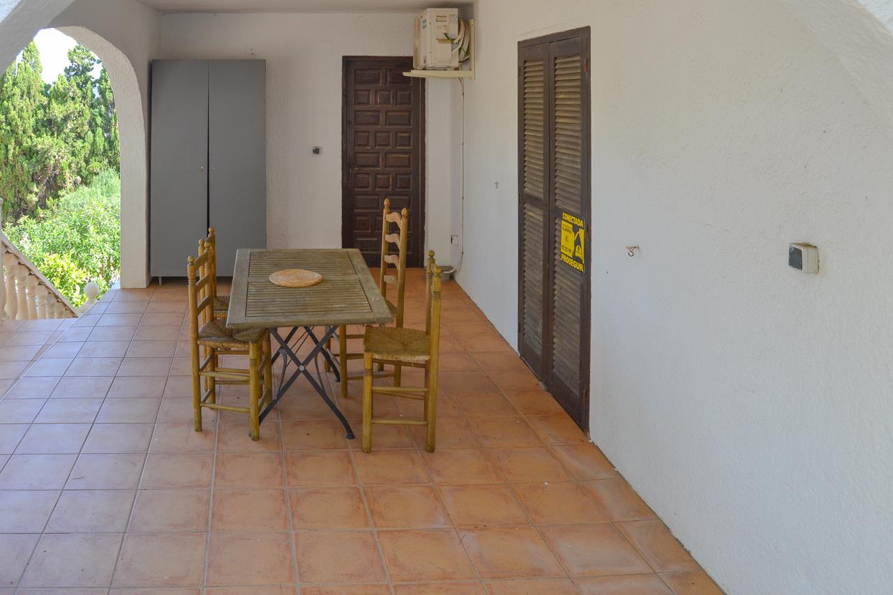 Terrasse couverte avec sol en terre cuite, comprenant une table à manger en bois et des chaises rustiques, bordée d'arches donnant sur le jardin.