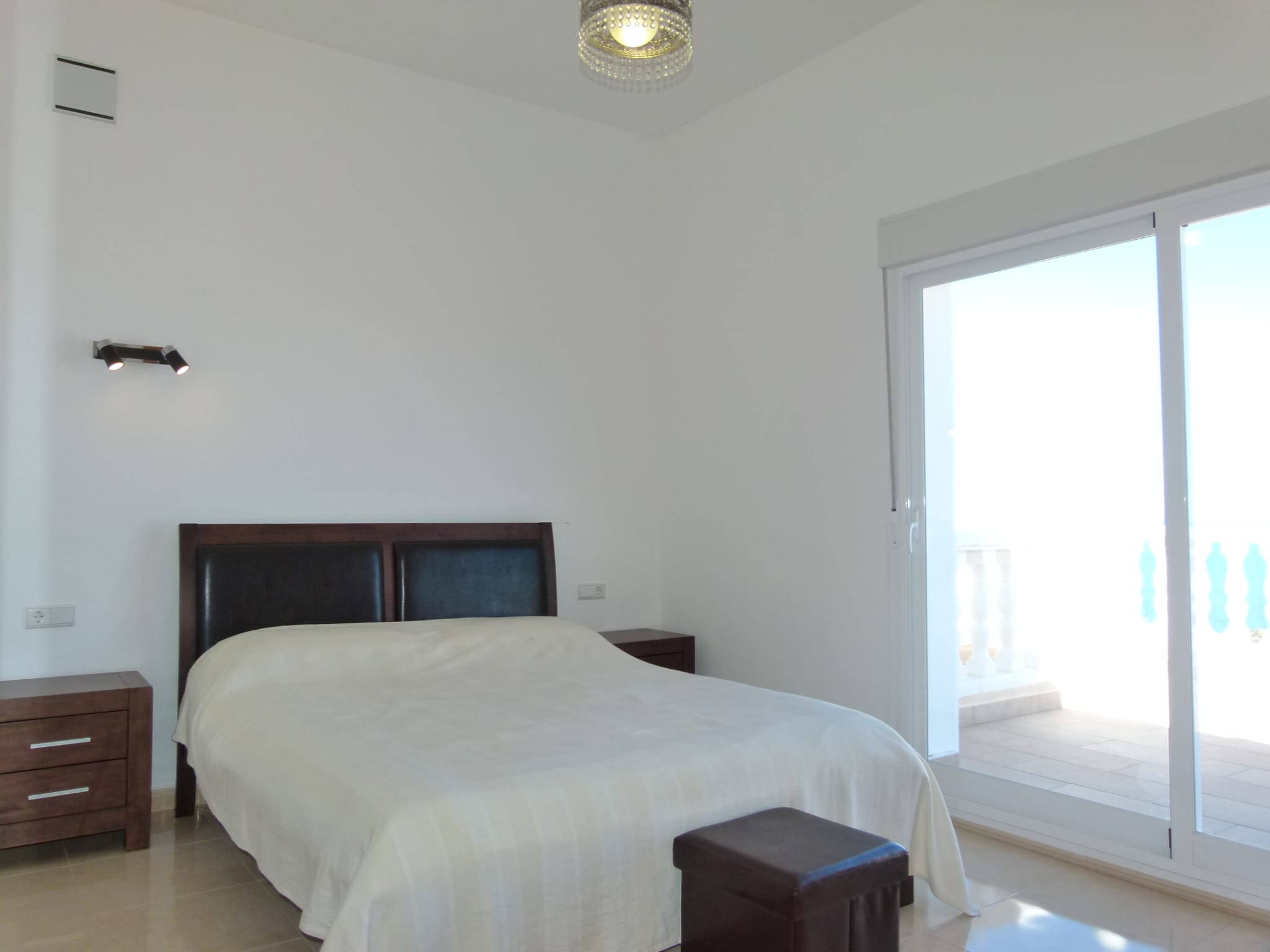 Chambre lumineuse avec lit double, tables de chevet en bois foncé et accès à une terrasse avec rambarde blanche.