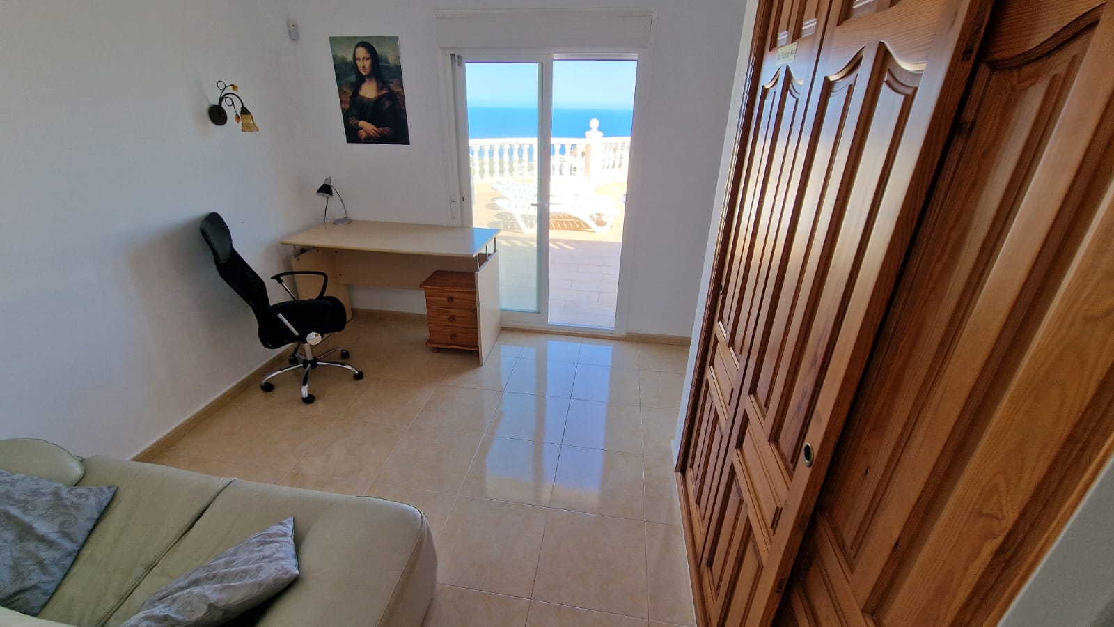 Pièce lumineuse avec bureau, armoire en bois et accès à la terrasse avec vue sur la mer.