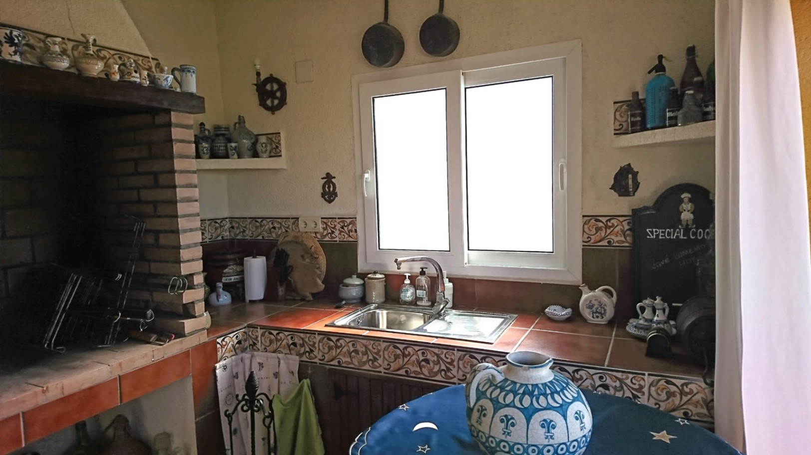 Cuisine d'été avec barbecue en briques, plan de travail en carrelage traditionnel, évier en inox et fenêtre en PVC offrant une luminosité naturelle.