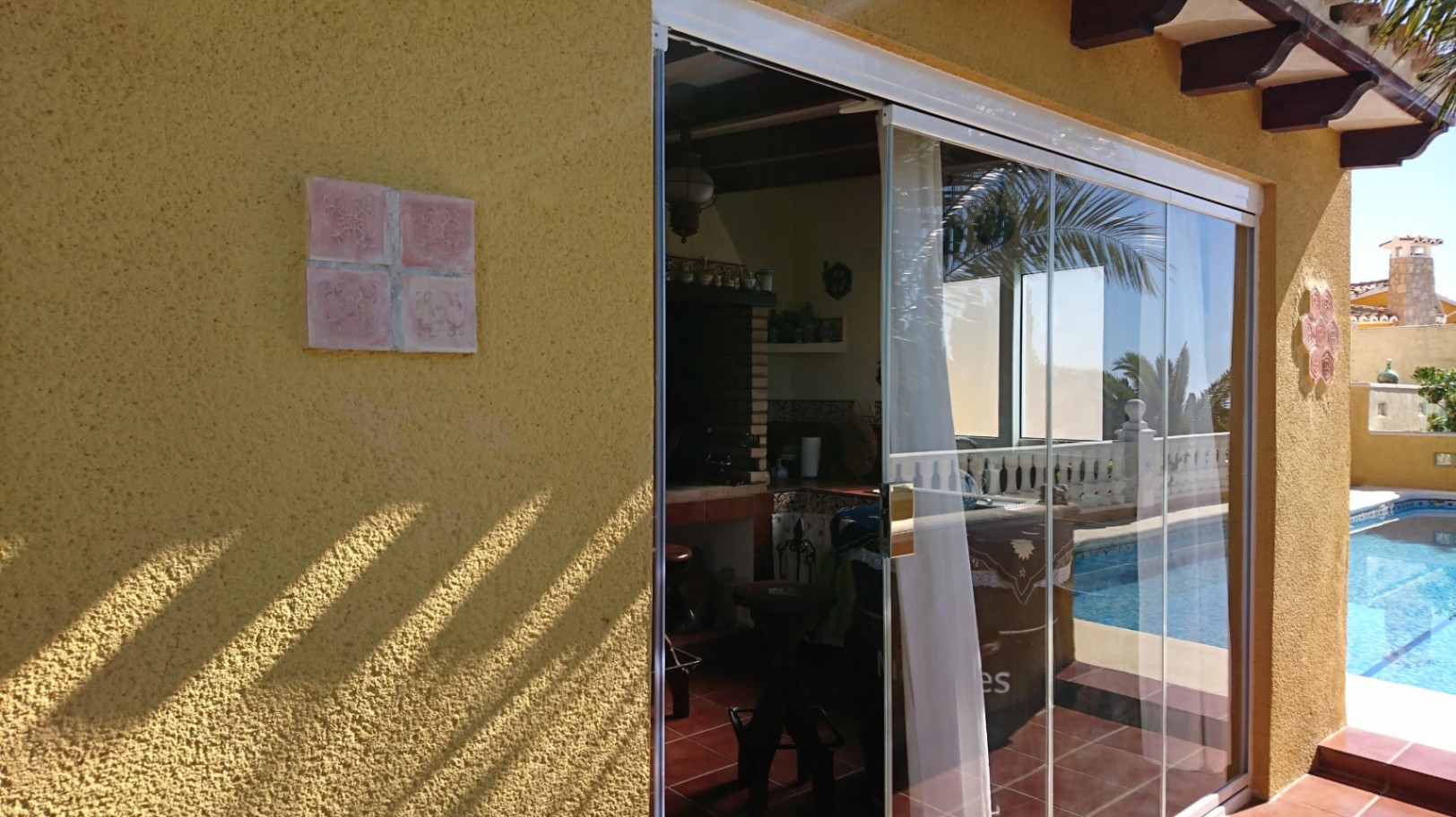 Cuisine d'été fermée par des rideaux de verre, avec barbecue en briques, poutres en bois et accès direct à la terrasse de la piscine.