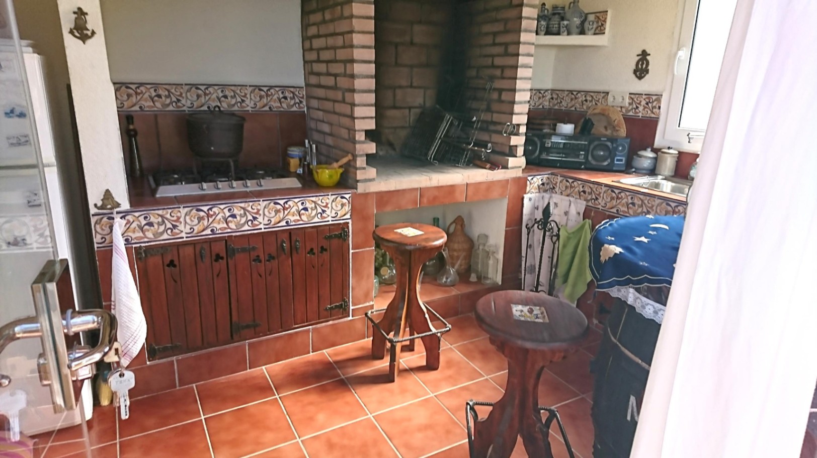 Cuisine d'été rustique avec barbecue en briques, plans de travail en carreaux décoratifs, meubles en bois et sol en terre cuite traditionnelle.