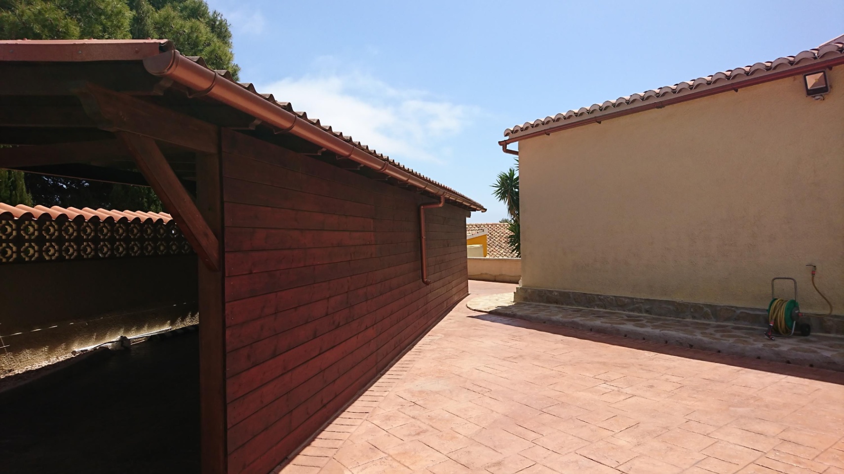 Espace extérieur pavé comprenant un abri en bois et la façade de la villa, tous deux surmontés de toitures en tuiles romaines traditionnelles.