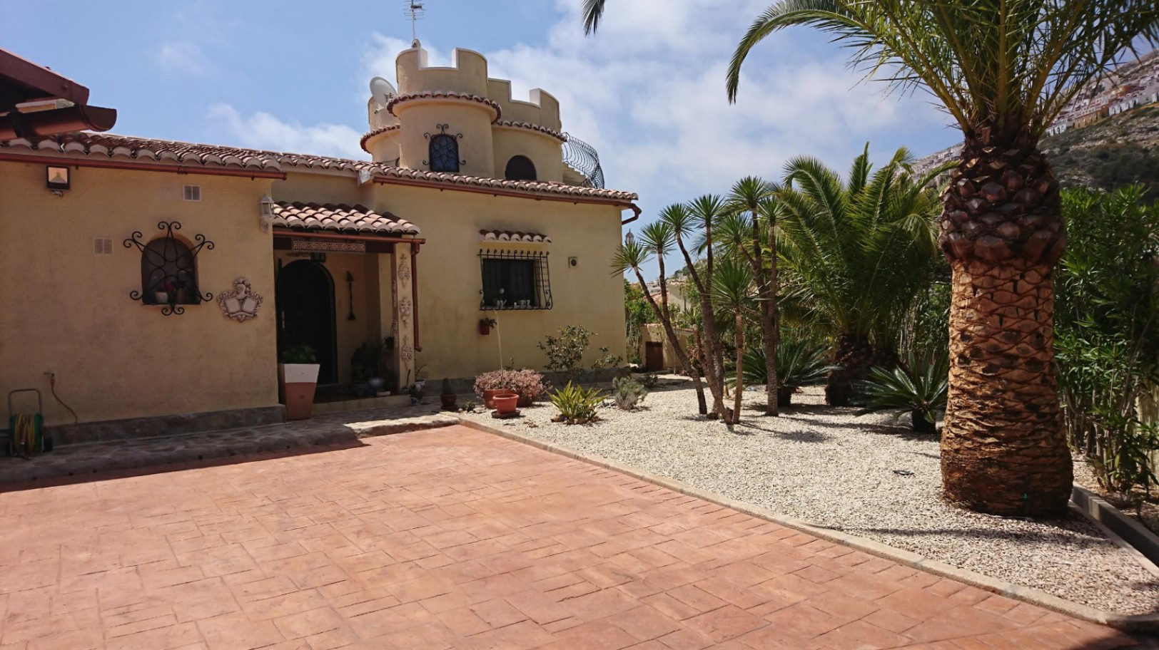 Extérieur de villa méditerranéenne avec tour crénelée, allée en terre cuite et jardin de gravier orné de palmiers et de yuccas.
