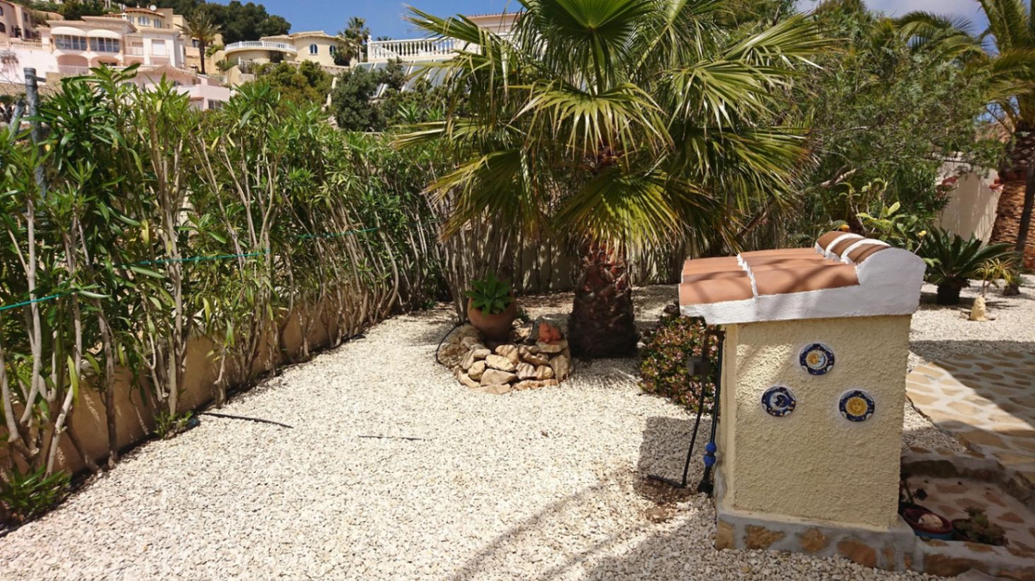 Jardin méditerranéen à faible entretien avec gravier blanc, palmiers et haies, agrémenté d'une structure décorative aux azulejos traditionnels.
