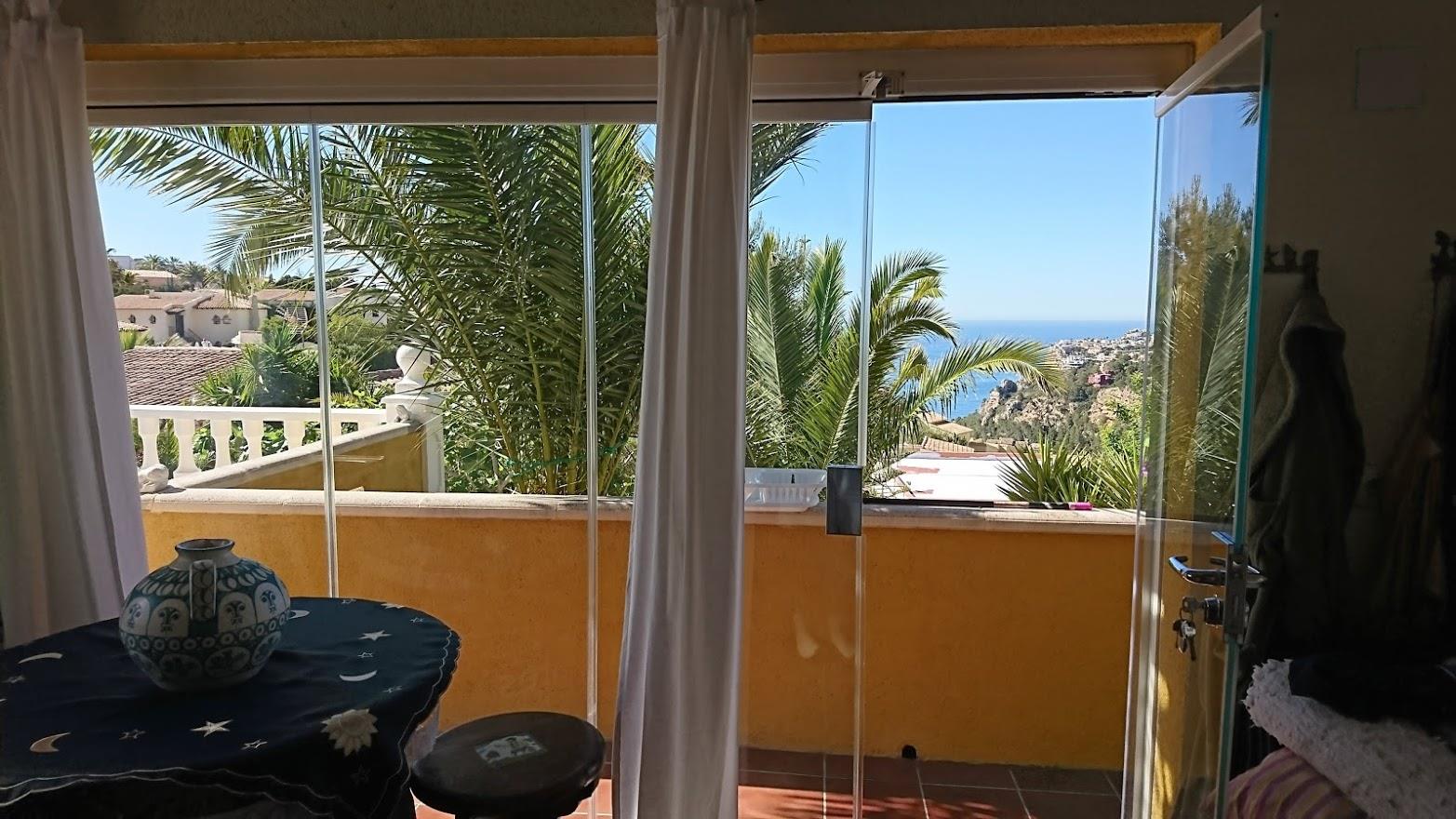 Naya vitrée offrant une vue sur la mer et les palmiers. Les rideaux de verre s'ouvrent sur un balcon avec balustrade, inondant l'espace de lumière naturelle.