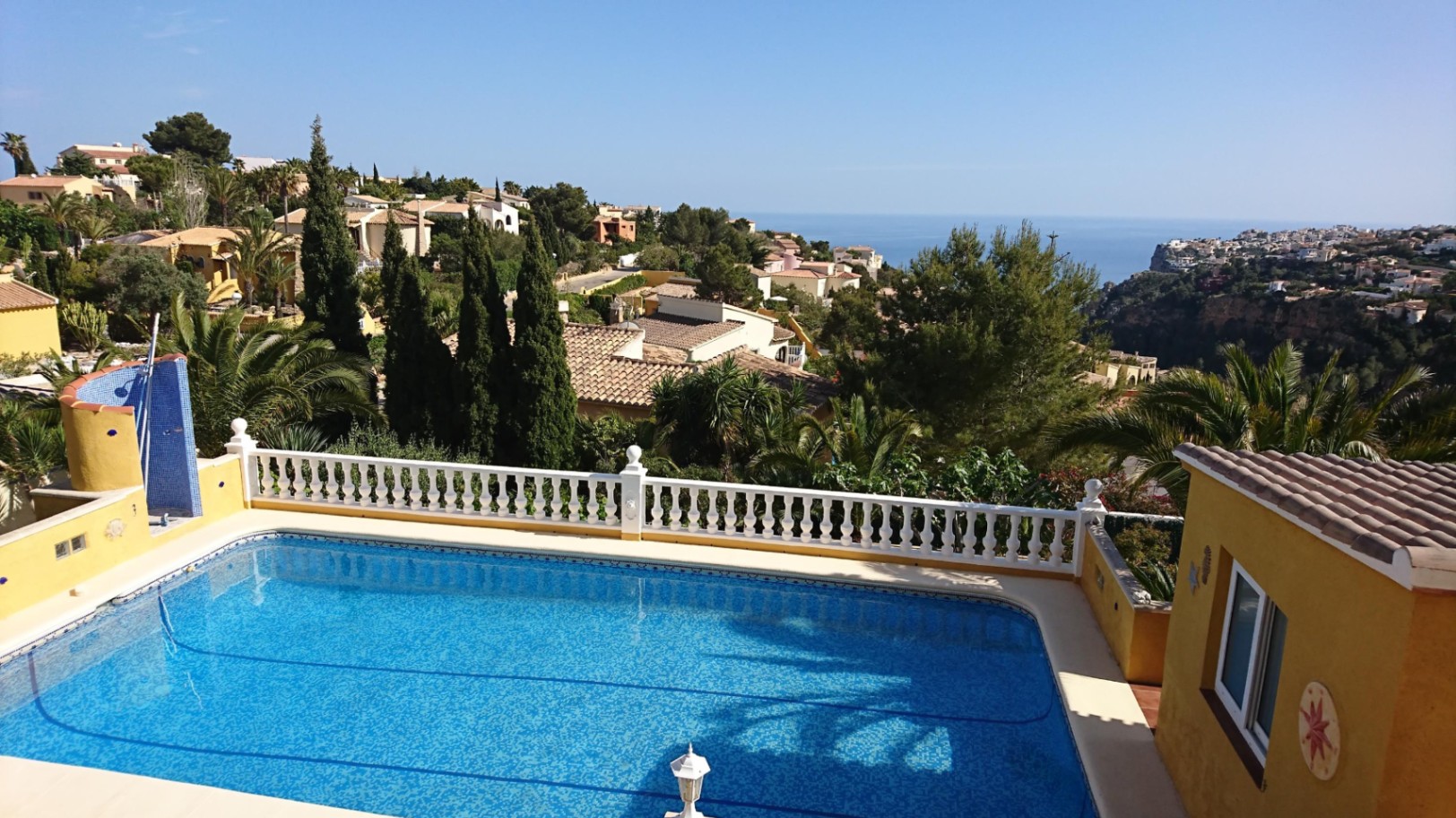 Piscine rectangulaire avec balustrade blanche et douche extérieure, offrant une vue dégagée sur la mer Méditerranée et les collines environnantes.