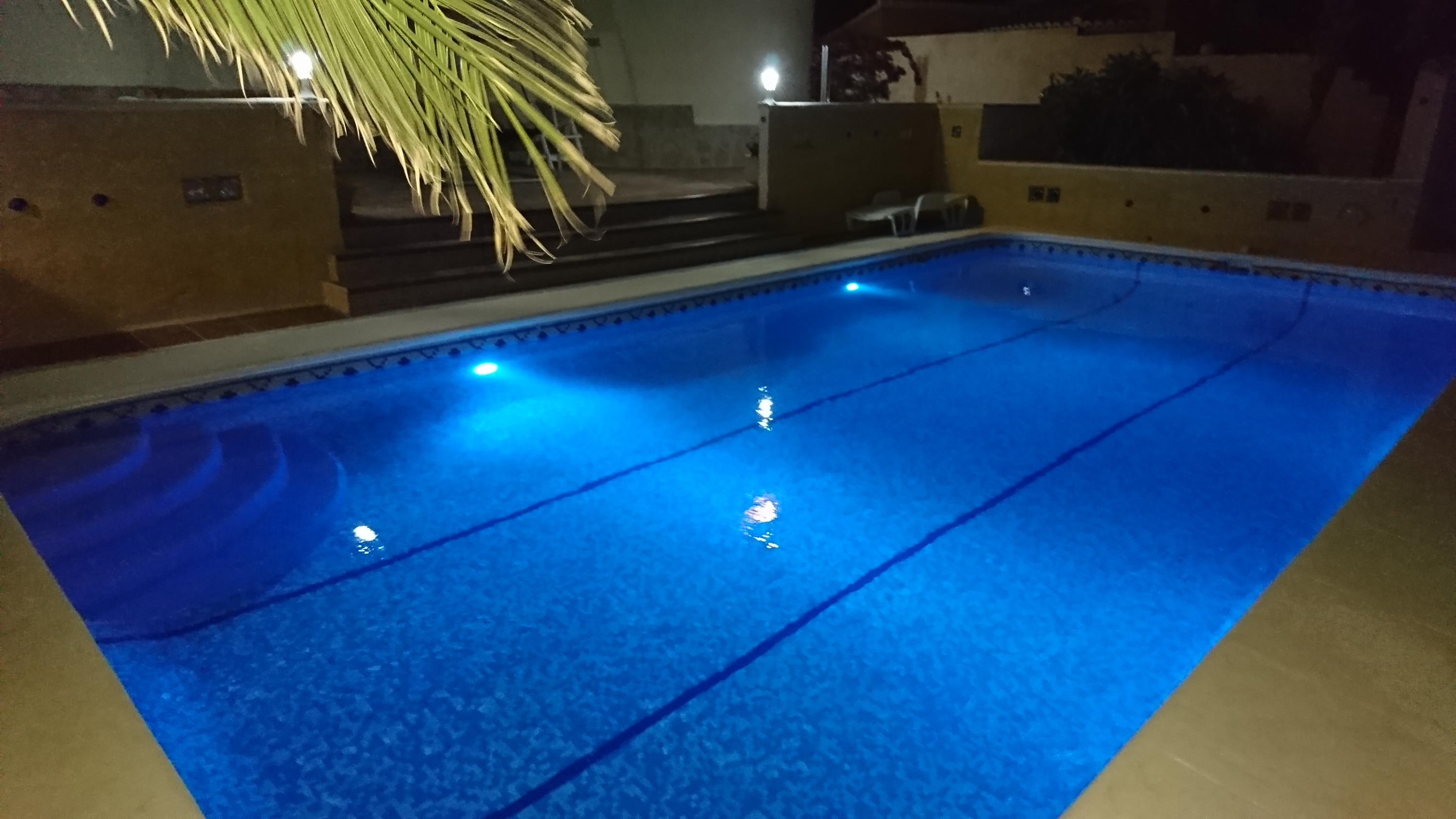Piscine rectangulaire avec éclairage subaquatique bleu, escalier romain et terrasse pavée sous les lumières du soir.