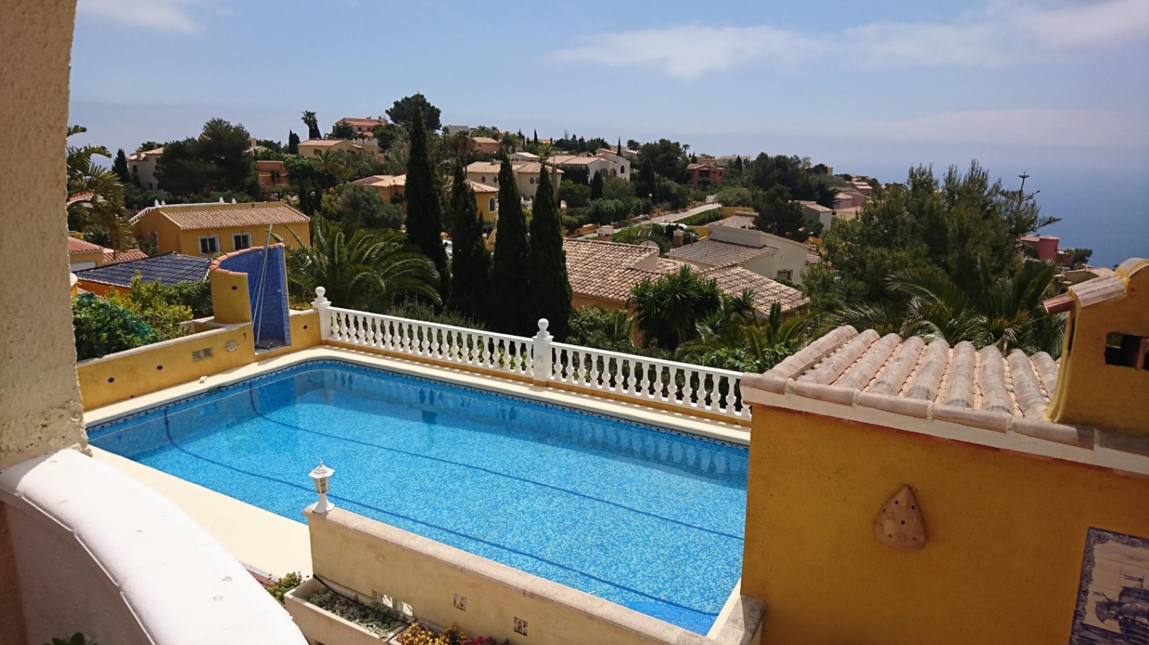 Piscine rectangulaire de 10x4 en mosaïque bleue, bordée d'une balustrade blanche offrant une vue dégagée sur la mer et le paysage résidentiel environnant.