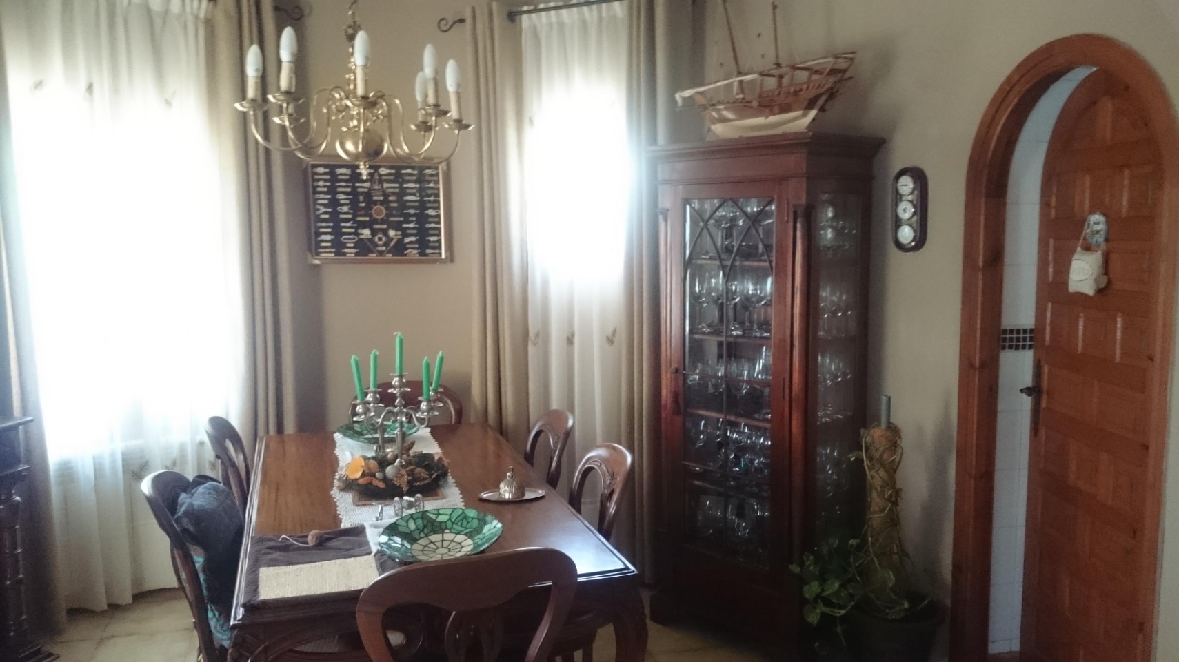 Salle à manger classique avec table en bois, vitrine d'exposition et lustre en laiton. Une porte cintrée en bois complète cet espace baigné de lumière.