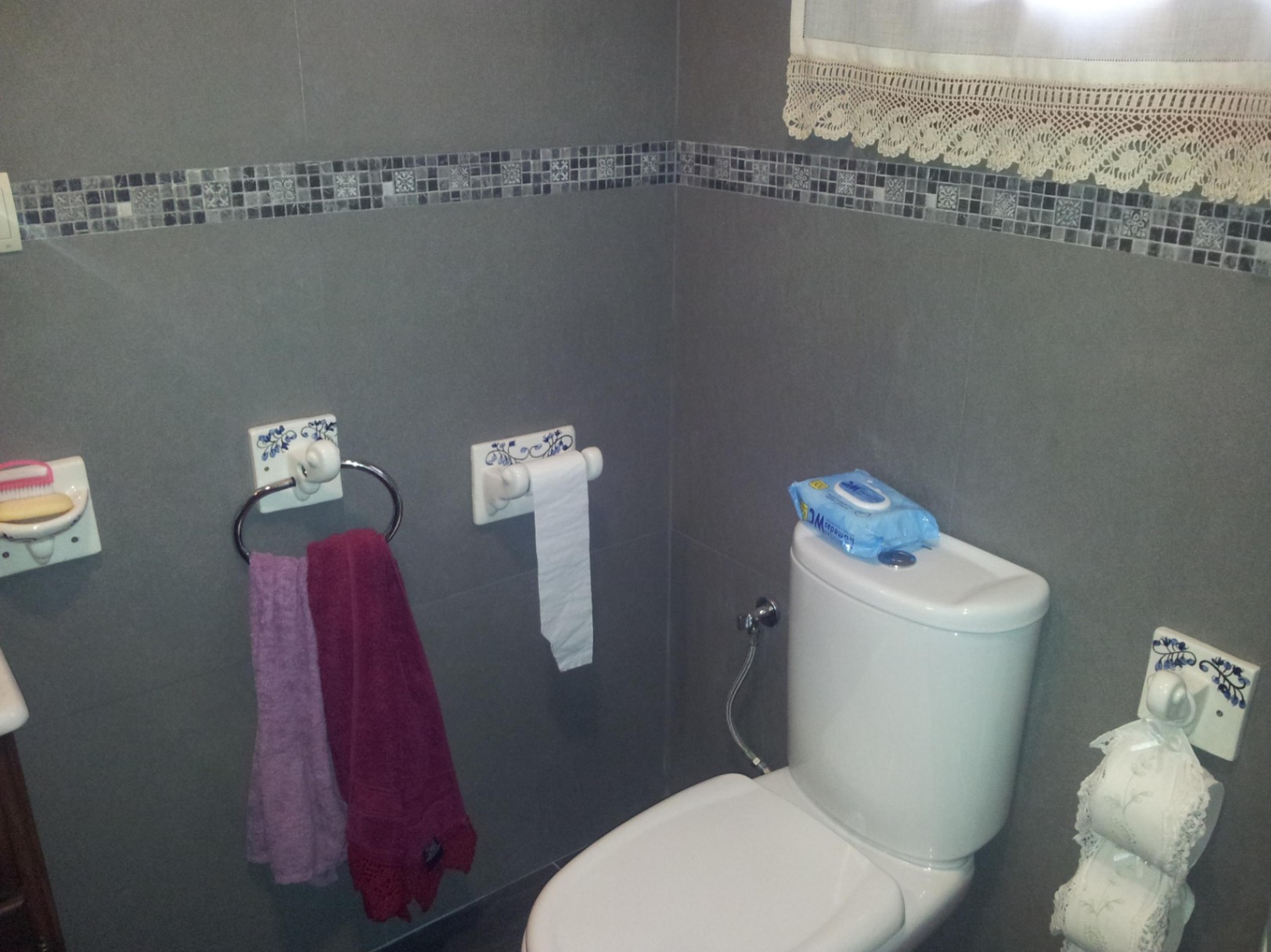 Salle de bain avec carrelage gris et frise en mosaïque, comprenant des sanitaires blancs et des accessoires en céramique aux motifs floraux bleus.