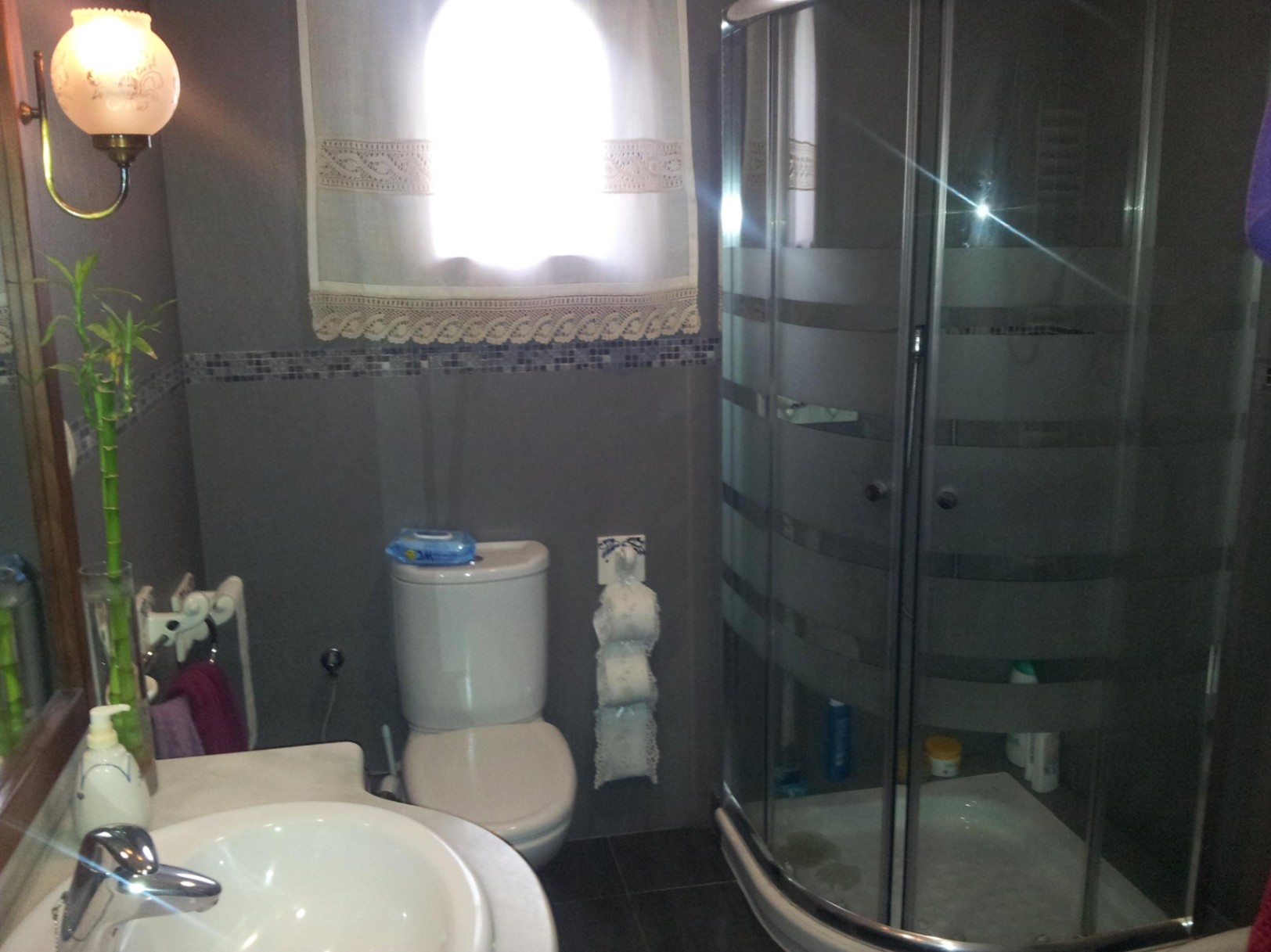 Salle de bain équipée d'une douche d'angle en verre courbé, de murs carrelés gris avec frise en mosaïque et d'un lavabo blanc.