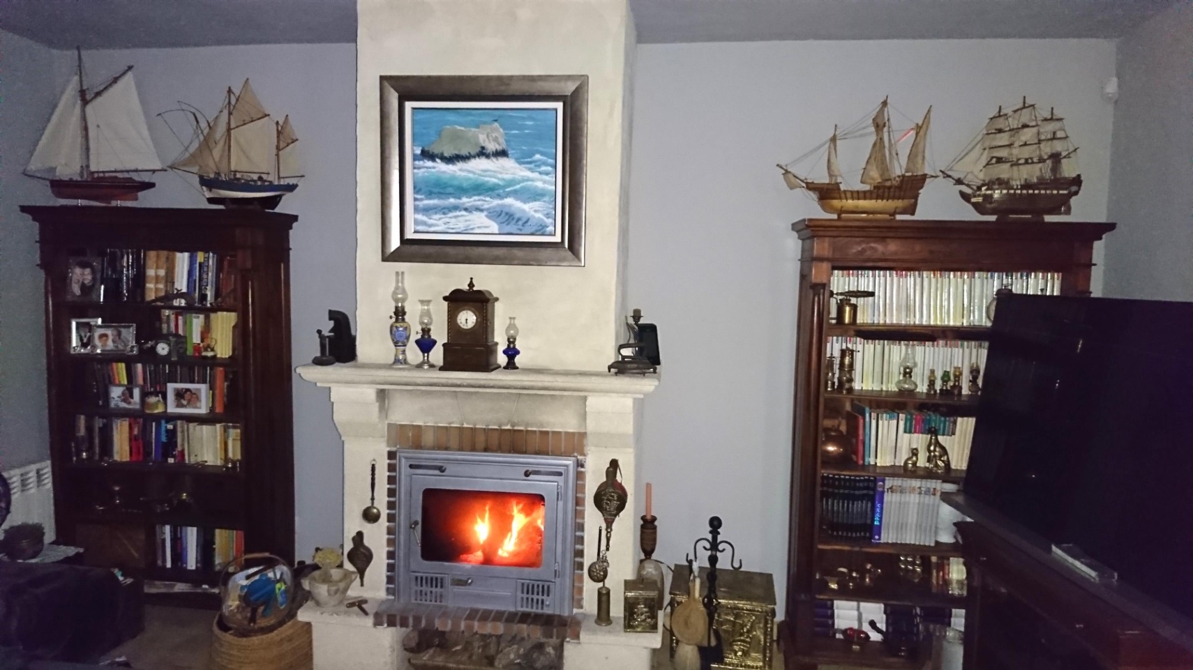 Salon avec cheminée en pierre équipée d'un insert, entourée de bibliothèques en bois massif ornées de maquettes de bateaux et d'objets anciens.