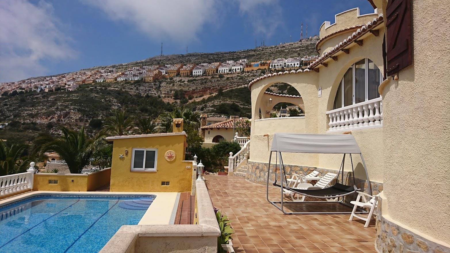 Terrasse avec piscine et mobilier de jardin devant la façade méditerranéenne, caractérisée par ses arches et sa tour, avec vue sur les collines environnantes.