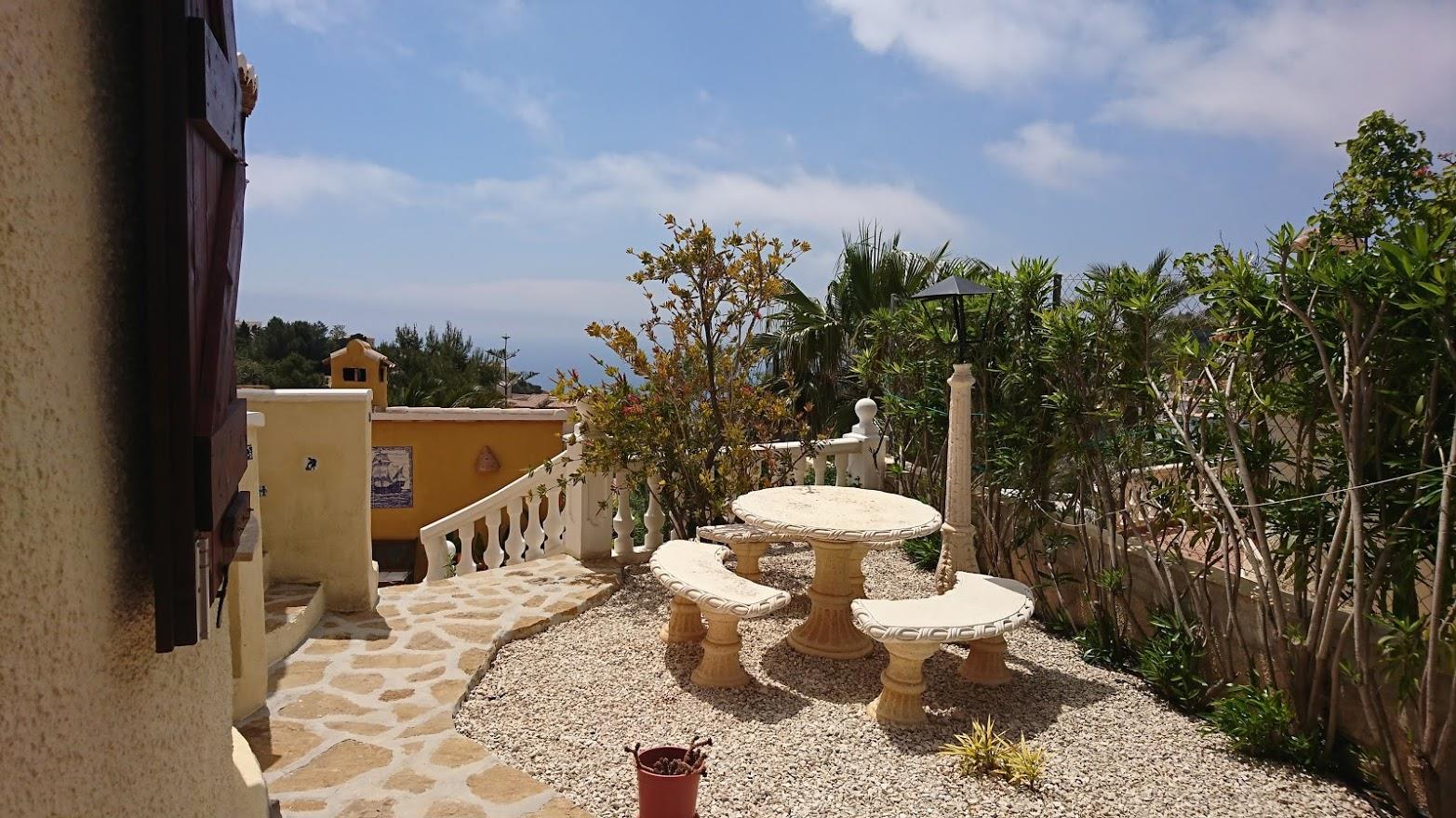 Terrasse en gravier avec table et bancs en pierre sculptée, bordée d'une balustrade blanche et de plantes méditerranéennes face à la mer.