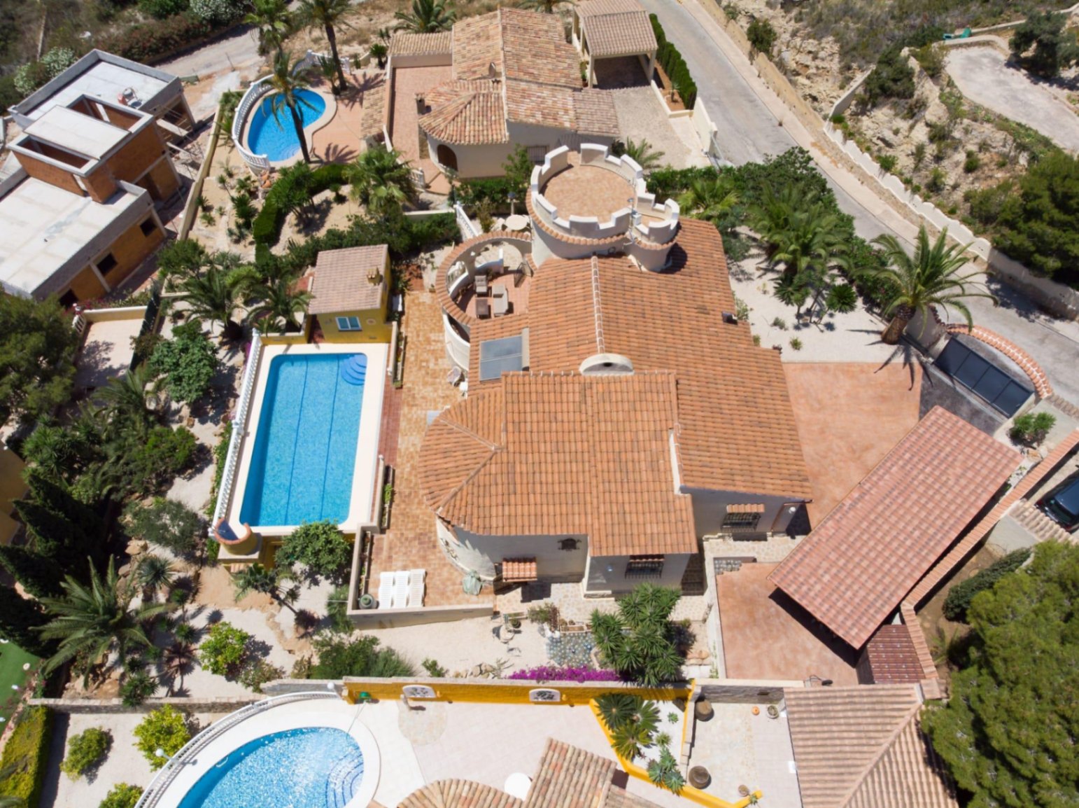 Vue aérienne de la villa montrant son toit en tuiles, sa tour circulaire crénelée unique et sa piscine rectangulaire entourée de palmiers et de jardins.