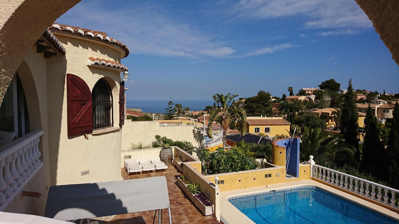 Vue extérieure avec piscine rectangulaire, terrasse en terre cuite avec transats et tour de la villa avec volets en bois et vue sur la mer.