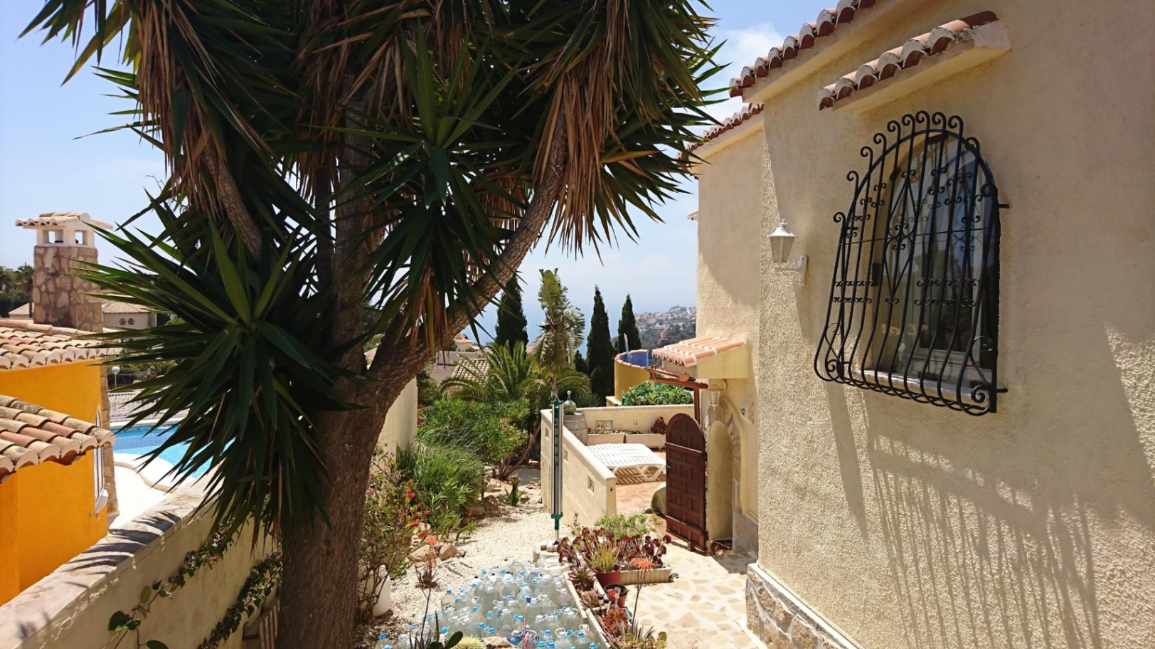 Vue extérieure de la villa avec façade en crépi, fenêtre ornée d'une grille en fer forgé et jardin méditerranéen offrant un aperçu sur la mer.