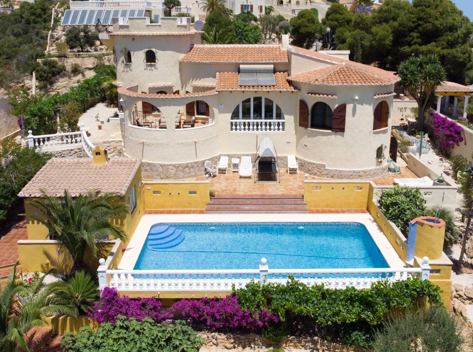 Vue extérieure de la villa avec piscine rectangulaire, terrasse en terre cuite et façade dotée d'une tour, entourée de végétation méditerranéenne et de murs jaunes.