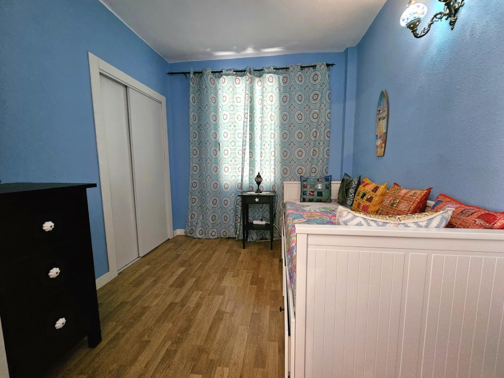 Chambre aux murs bleus avec parquet, placard intégré blanc et un lit gigogne agrémenté de coussins colorés à côté d'une commode noire.