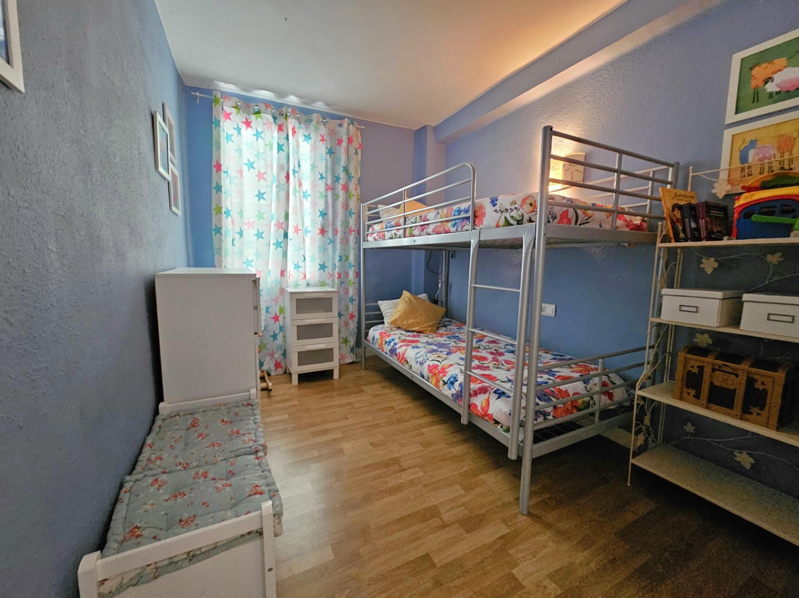 Chambre avec lits superposés en métal, parquet au sol et murs bleus. Elle dispose de rangements blancs et d'une fenêtre avec des rideaux à motifs d'étoiles.