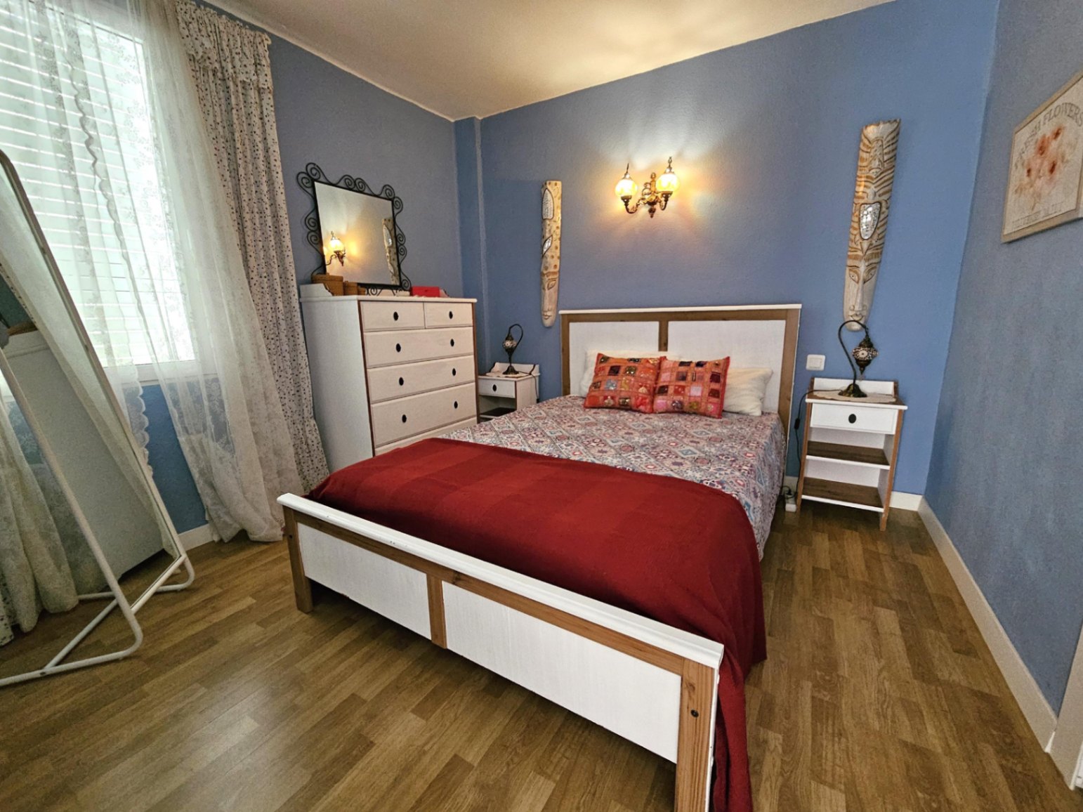 Chambre avec murs bleu clair, parquet et mobilier en bois blanc. Elle comprend une commode, un miroir sur pied et une lumière naturelle filtrée par des rideaux.