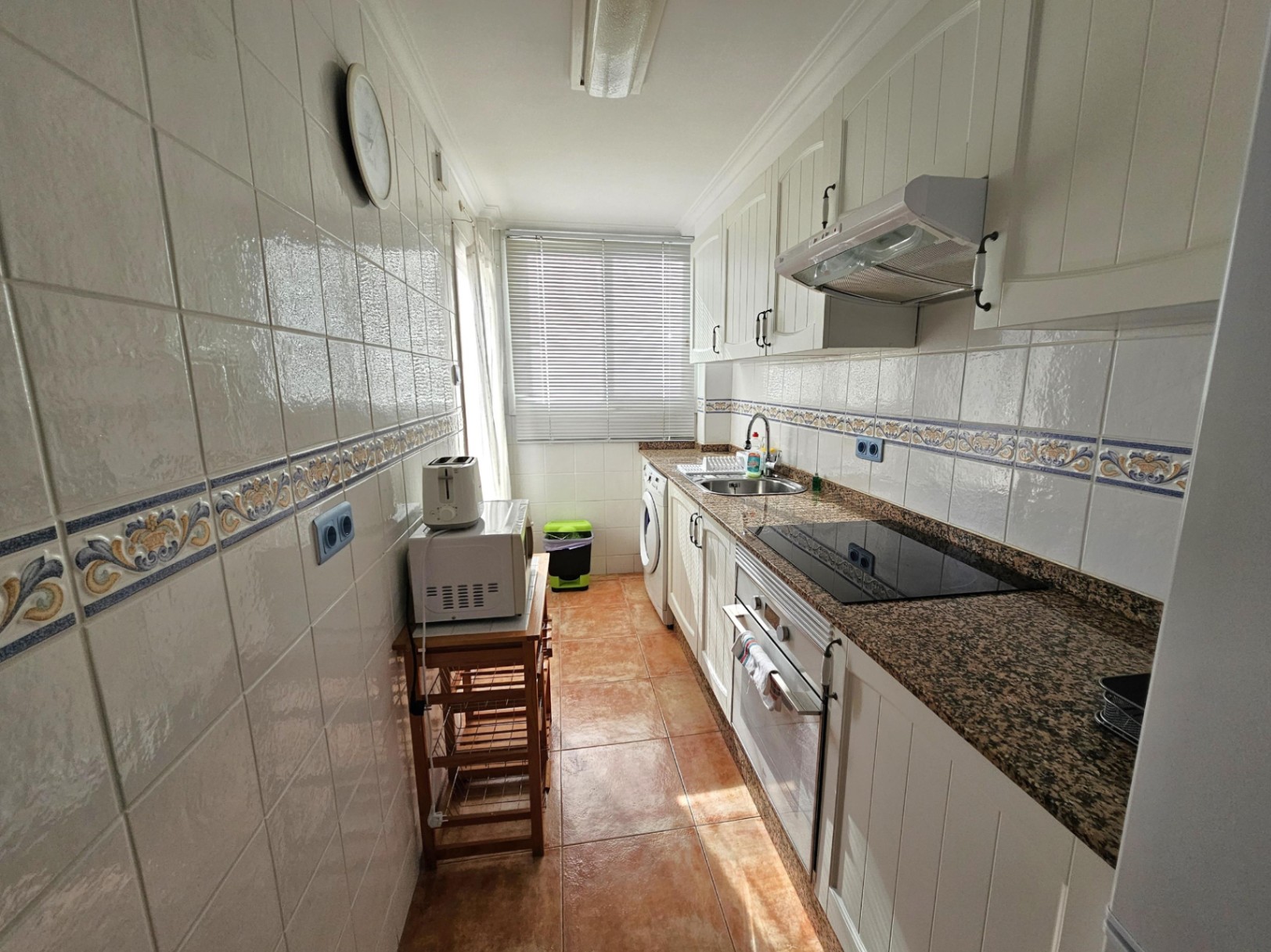 Cuisine indépendante en longueur avec plan de travail en granit, meubles blancs et électroménagers encastrés. Une fenêtre apporte une lumière naturelle sur l'espace buanderie.