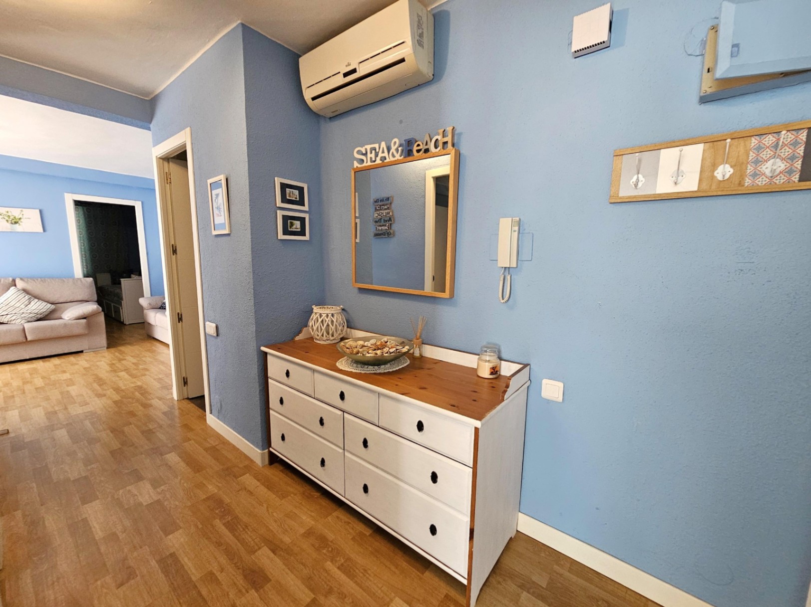 Entrée avec murs bleus, commode en bois blanc, parquet au sol et climatisation split, offrant une vue dégagée sur le salon attenant.