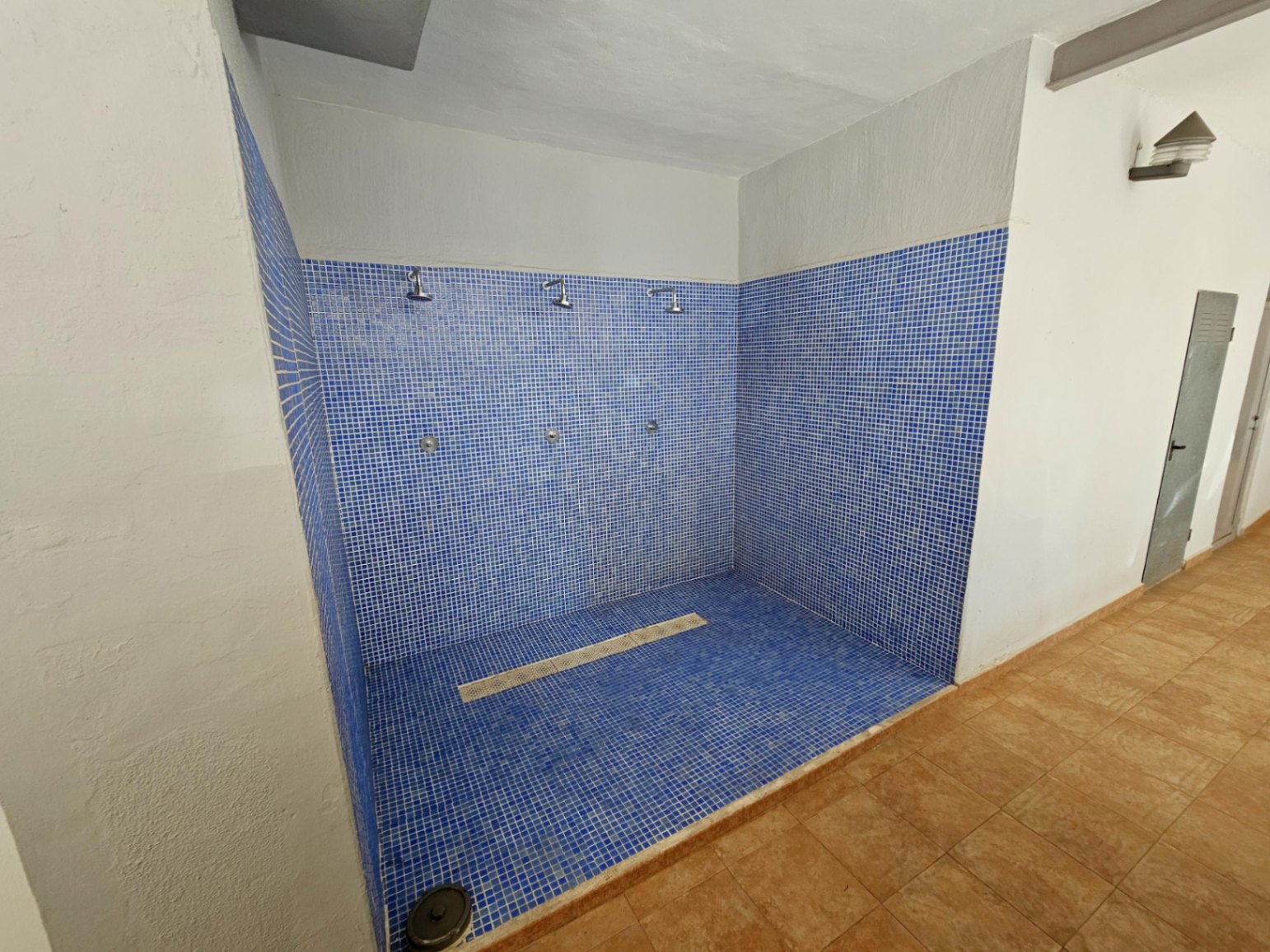 Espace de douches communes avec revêtement en mosaïque bleue, pratique pour se rincer après la plage. Comprend trois pommeaux de douche et un drainage linéaire au sol.