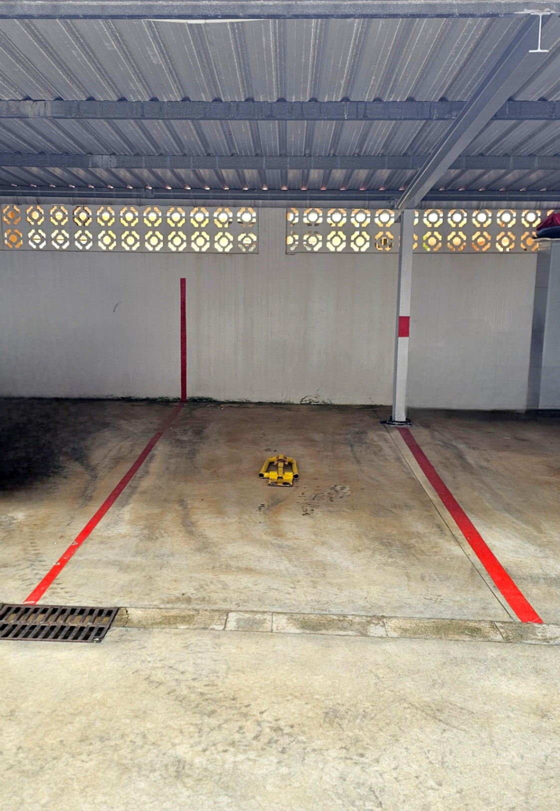 Place de parking privée et couverte avec toiture métallique, délimitée par des lignes rouges et équipée d'un arceau de sécurité jaune.