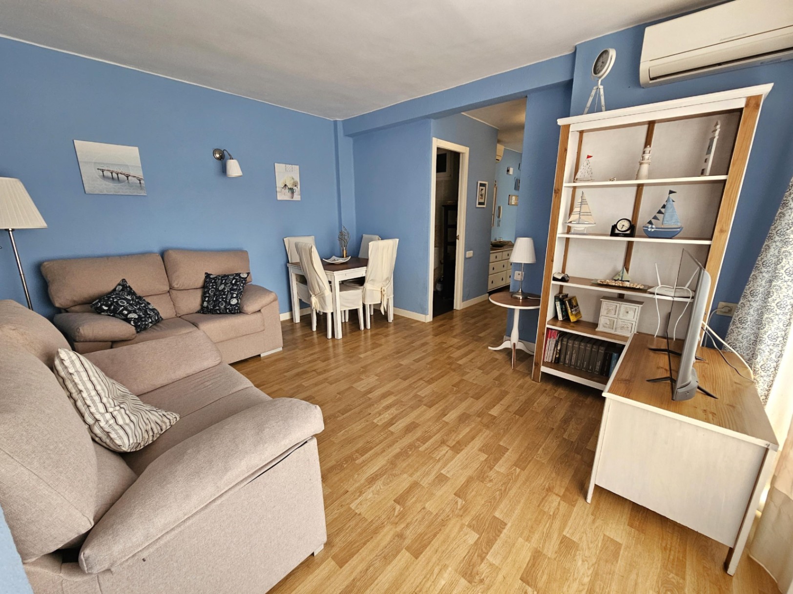 Salon et salle à manger avec murs bleus, parquet et canapé beige. Comprend une climatisation murale et un meuble de rangement blanc avec décoration nautique.